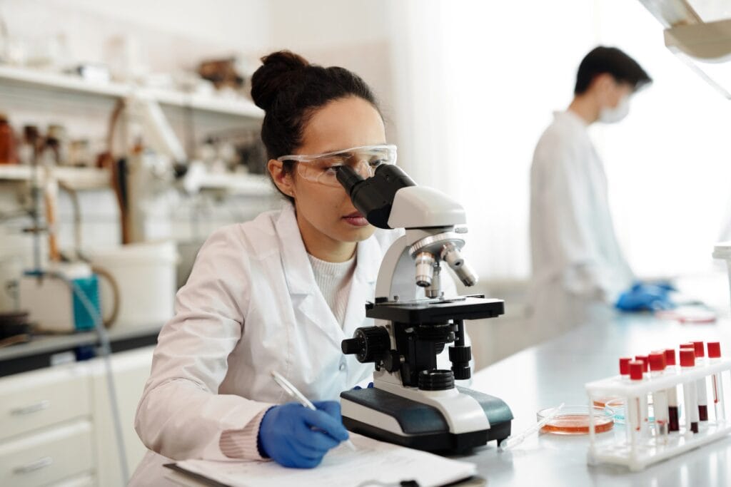 cientifica con bata de laboratorio blanca viendo a traves de un microscopio y tomando notas