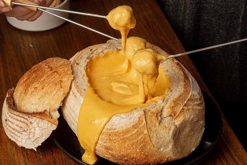 pan relleno de fondue de queso y dos personas remojan 2 trozos de comida en el fondue